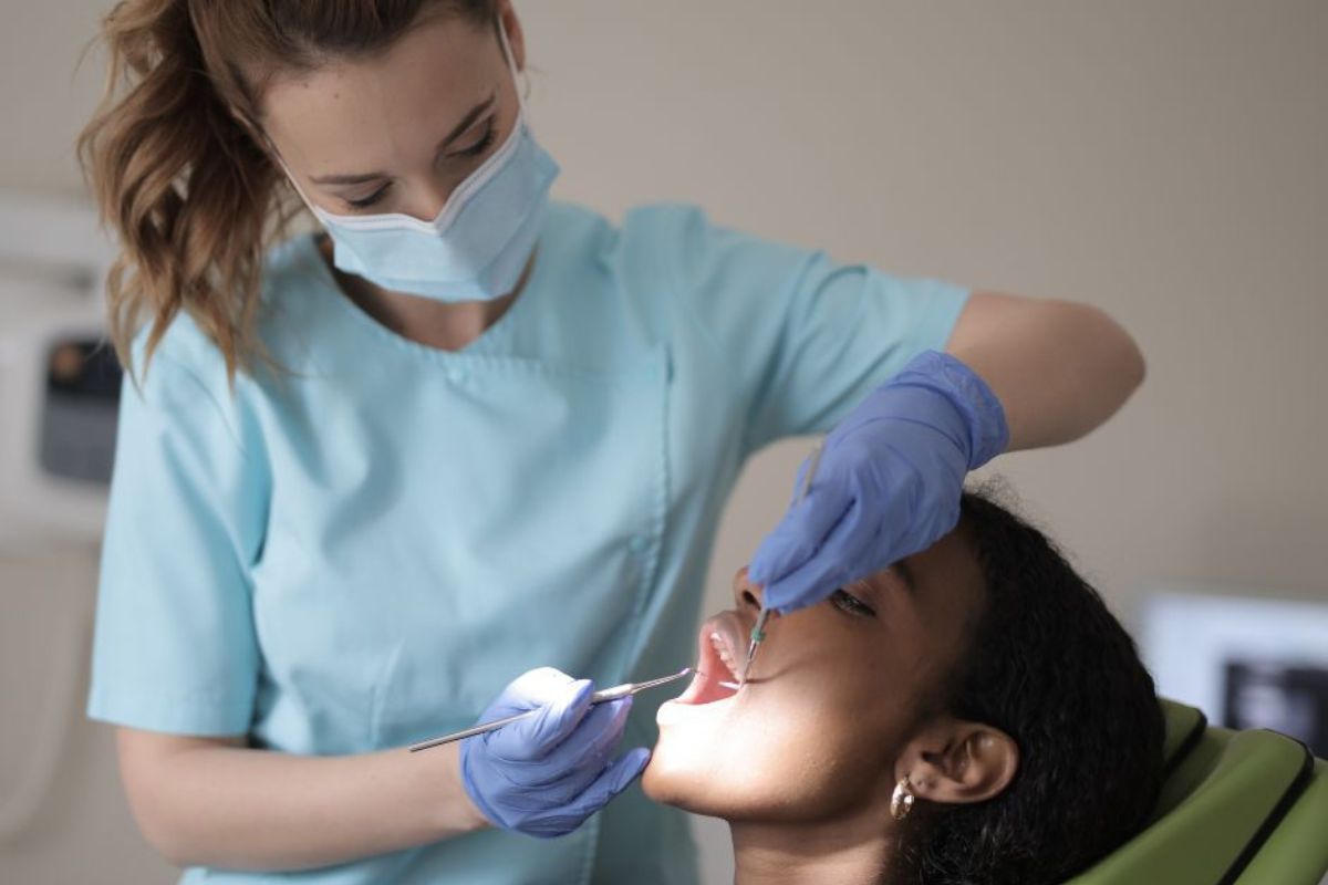 A young woman reclines in a dental chair while a masked and gloved dentist in blue scrubs conducts a dental examination using professional tools.