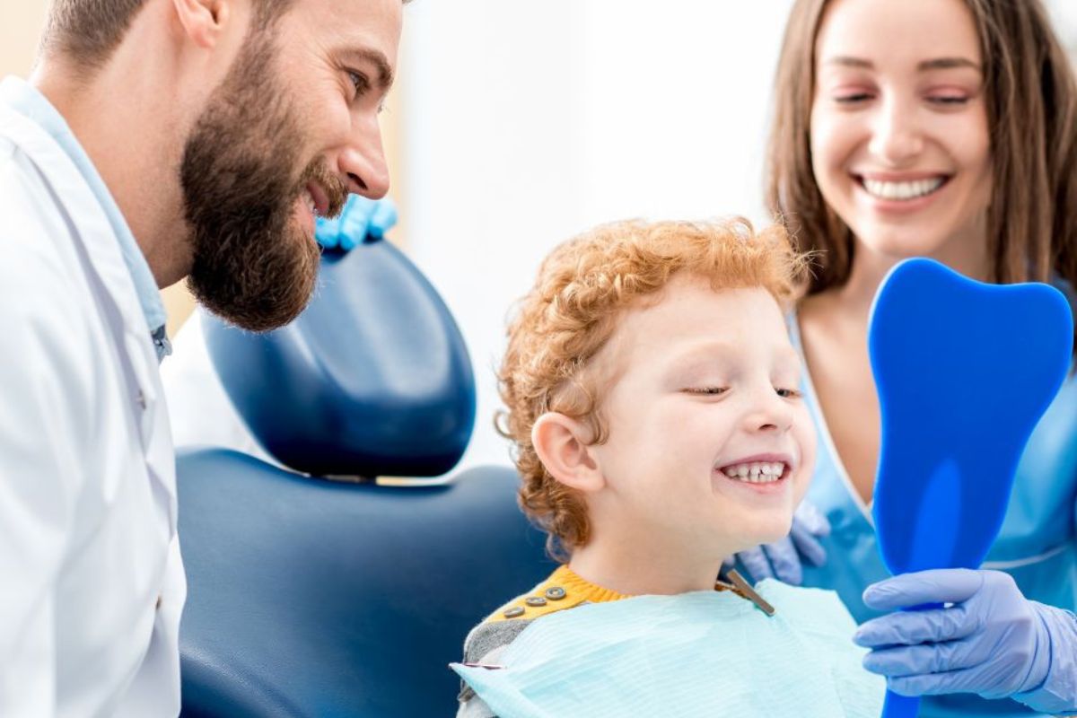 Smiling young boy looking at his teeth in a blue mirror after a dental check-up, with a happy dentist and dental assistant beside him.