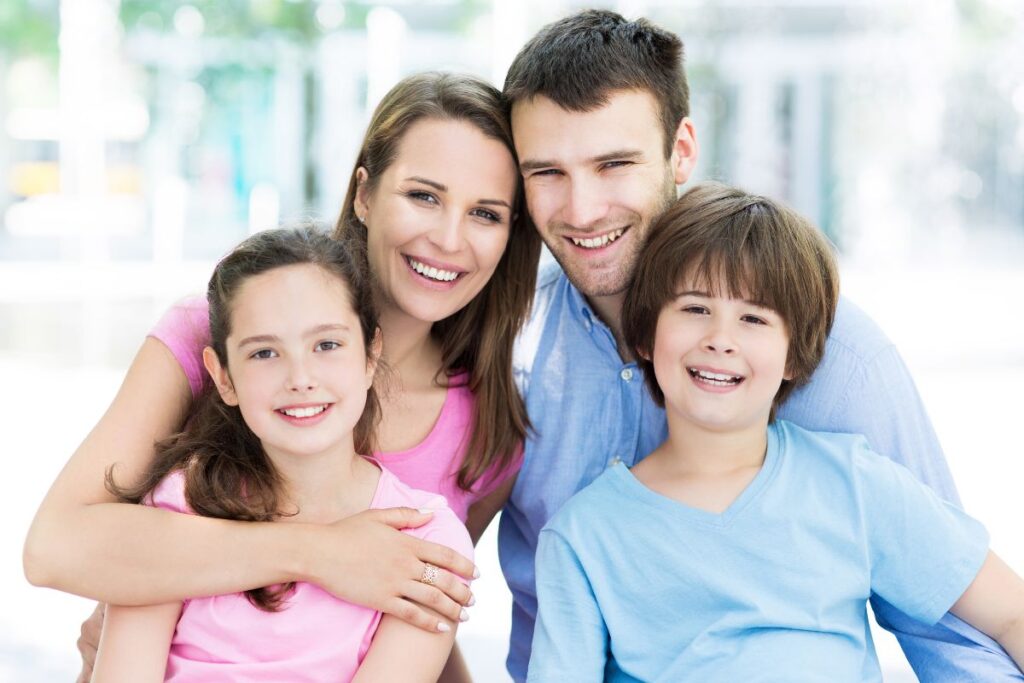 Smiling family of four, including two children, posing closely together in a bright, outdoor setting.