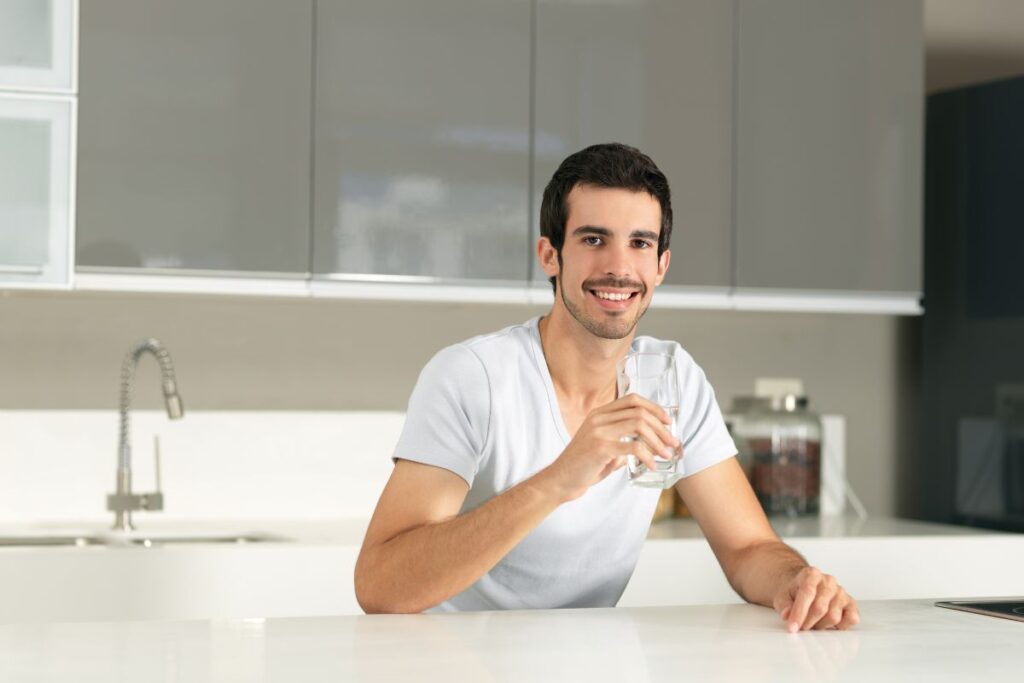 Smiling man in a kitchen holding a glass of water, promoting hydration and healthy lifestyle habits.