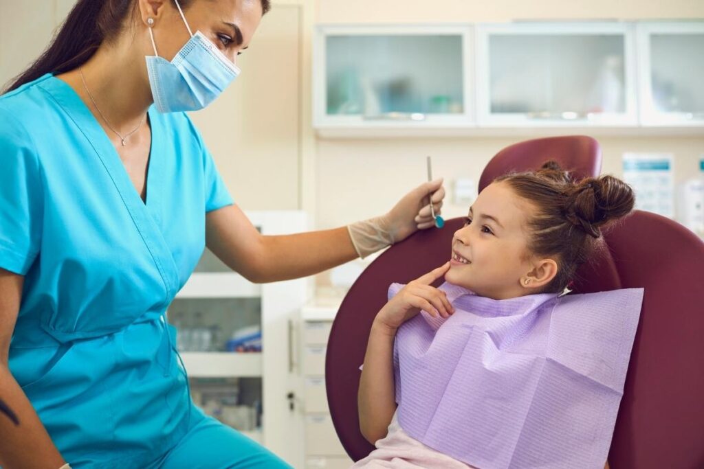 Young girl pointing at her teeth while a masked dentist prepares for examination in a bright dental clinic.