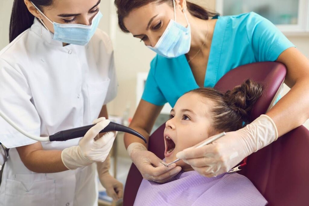 Dentist and assistant examining a young girl’s open mouth during a dental check-up, both wearing masks and gloves.