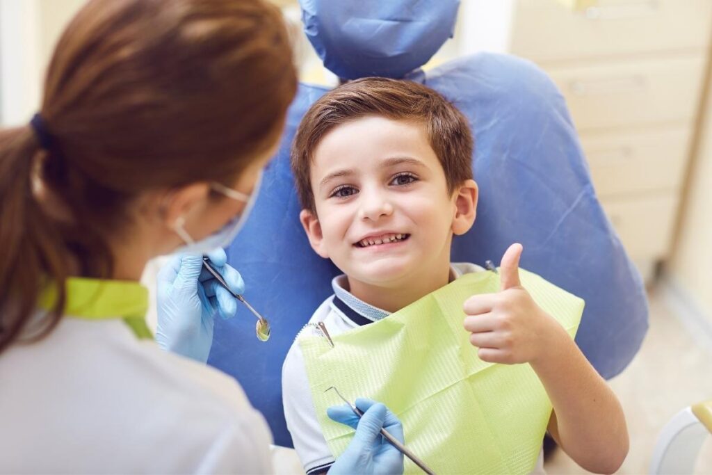 Smiling boy in dental chair giving a thumbs up while dentist in mask holds dental tools during check-up.