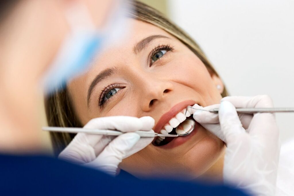 Close-up of a smiling patient at the dentist as gloved hands use a mirror and probe to examine her teeth.