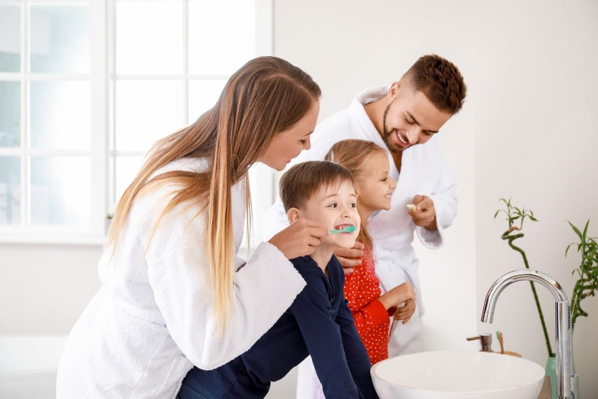 Family toothbrushing: mom guides boy at the vanity as dad and daughter smile and brush, all in white robes.