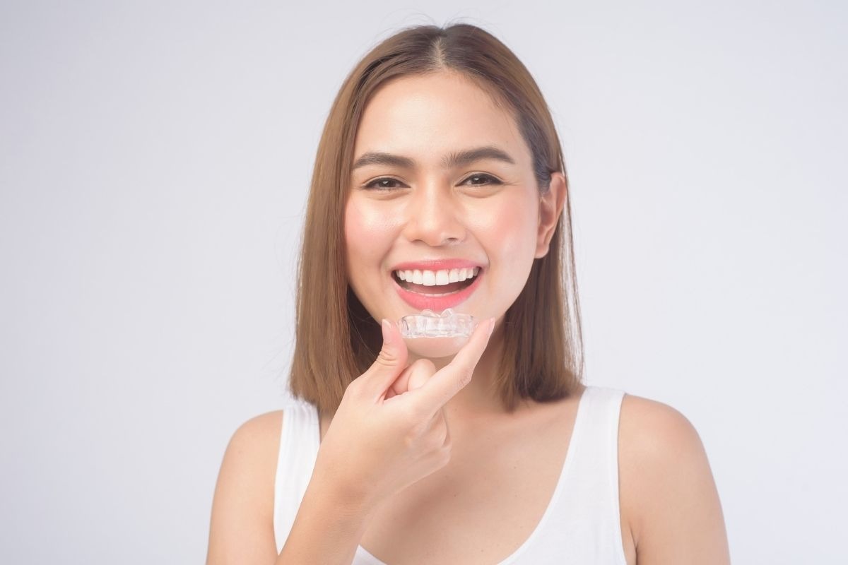 Smiling woman holds a clear dental aligner in front of her teeth, white tank top, light gray background.
