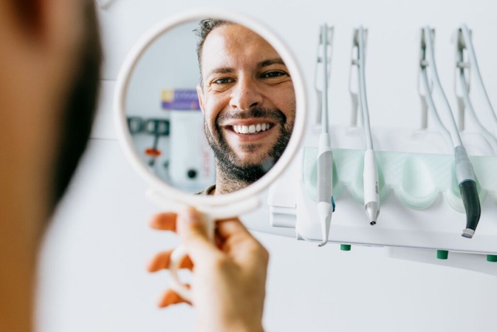 Man at dental clinic smiles into a round hand mirror; clean teeth and dental tools visible in the background.