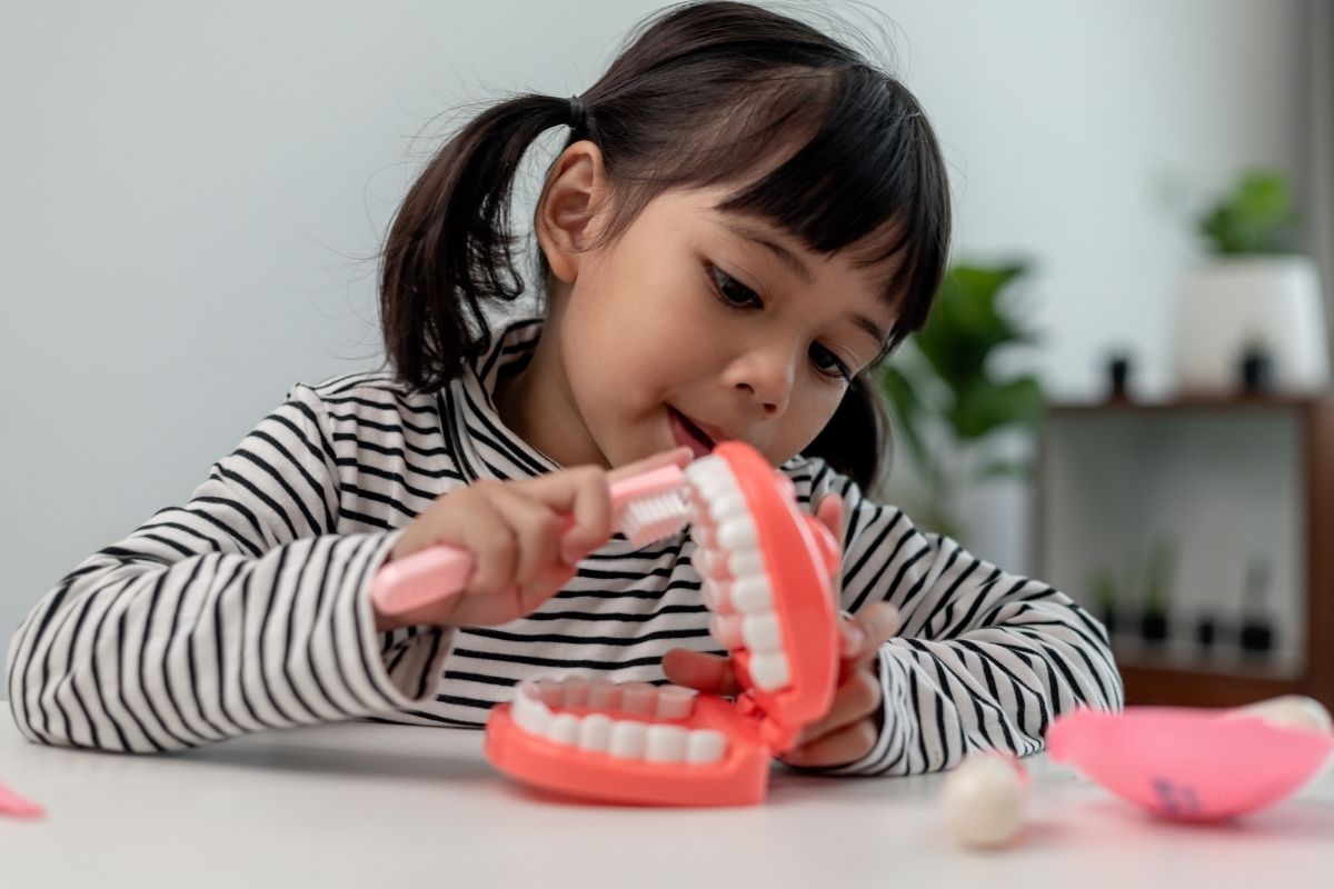 Little girl in a striped shirt practices brushing on a pink dental model with a toothbrush at a table.