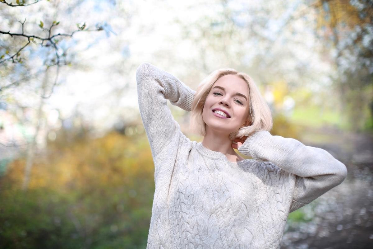 Smiling woman outdoors wearing a light sweater, standing on a nature path with soft sunlight and trees in the background.