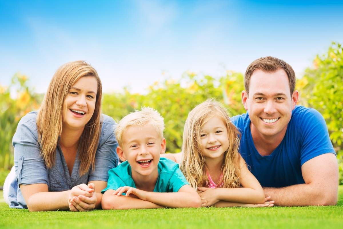 Happy parents and two children smiling while relaxing on a lawn with greenery in the background.